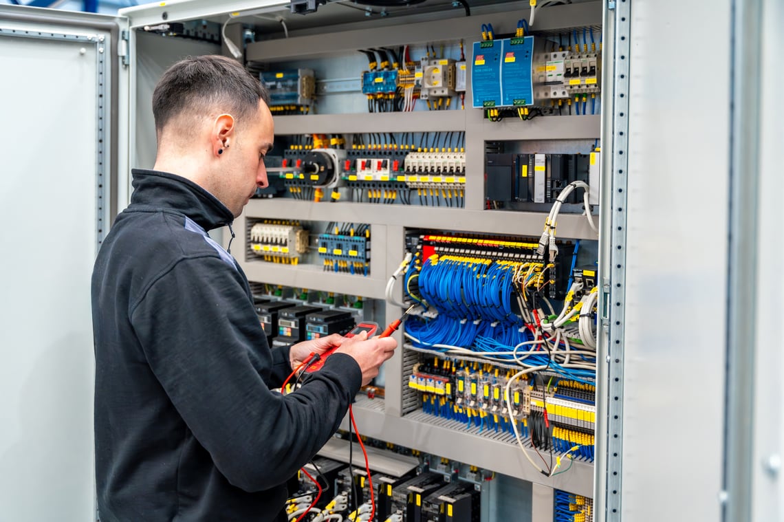 Electrician troubleshooting an open panel control cabinet with relays, terminal blocks, and labeled wiring.